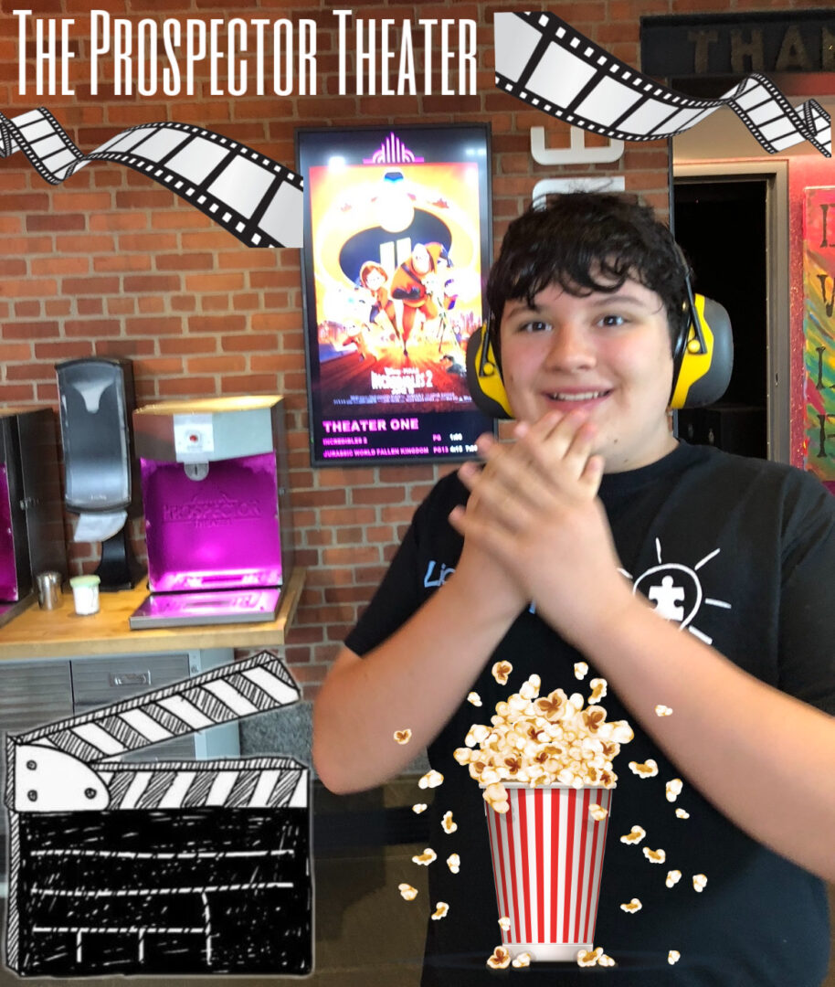 Mikey at the movies wearing his headphones and his light it up blue tee shirt, smiling in front of an Incredibles 2 poster