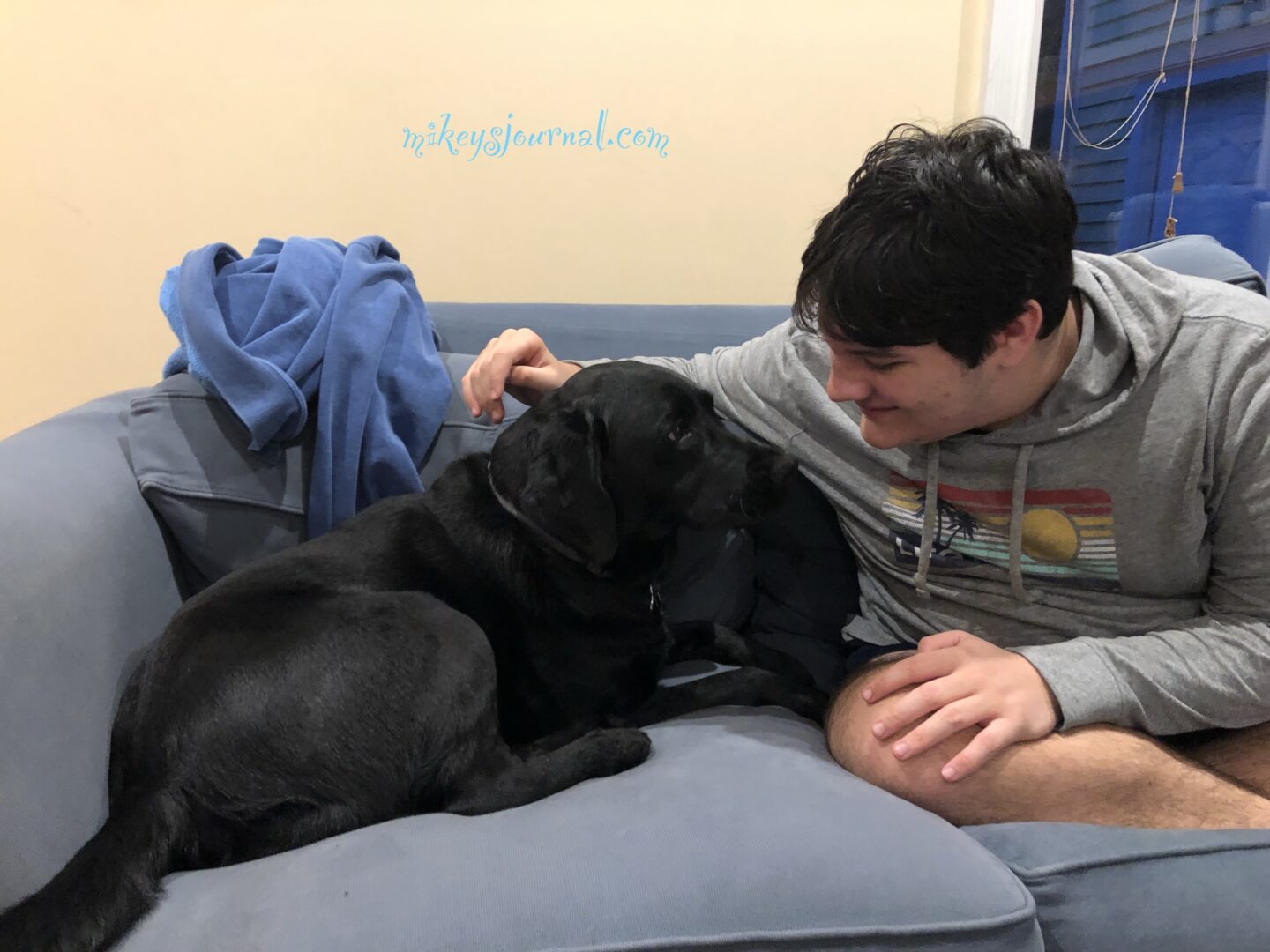Mikey sitting on the couch with our black lab Sonny. Mikey is leaning in and looking into Sonny’s eyes gently smiling.