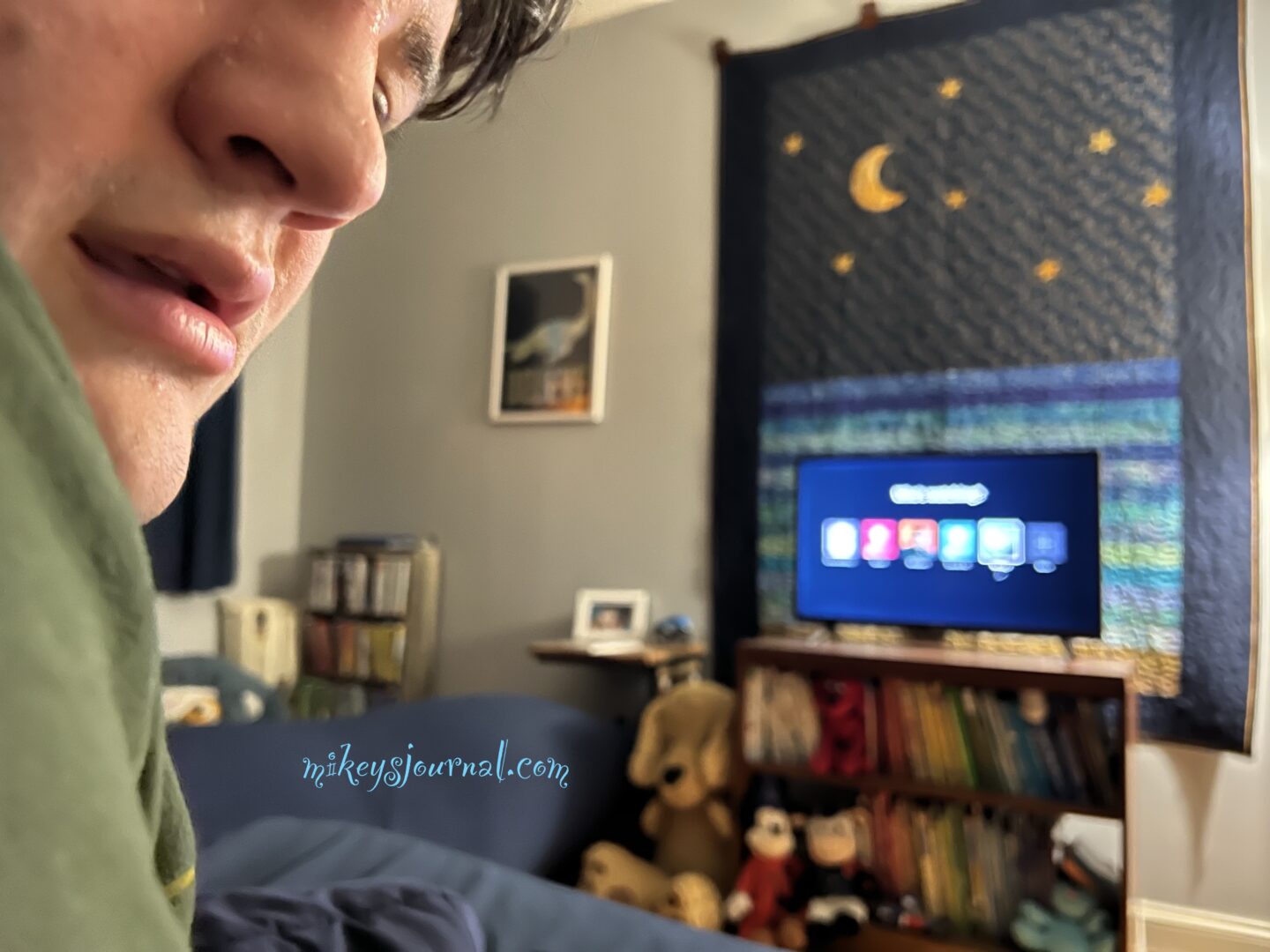Mikey, sitting in bed, looking at the camera with his TV on in the background.
