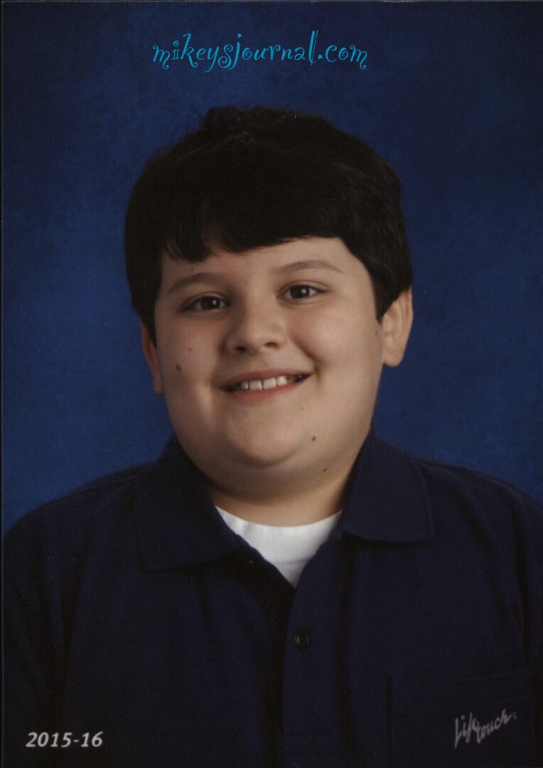Mikey’s 5th grade school photo, smiling, wearing a white tee under a navy polo shirt. 2015-16 and Lifetouch (the photo company) printed on the photo